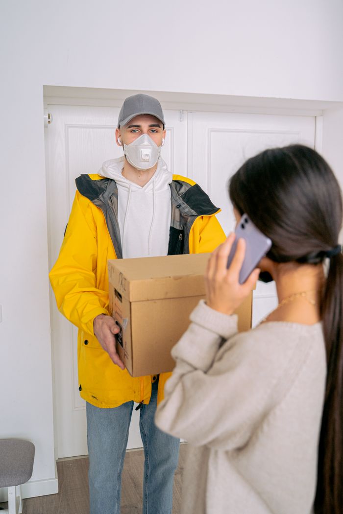 A delivery person hands a package to a woman inside a home, both wearing masks.